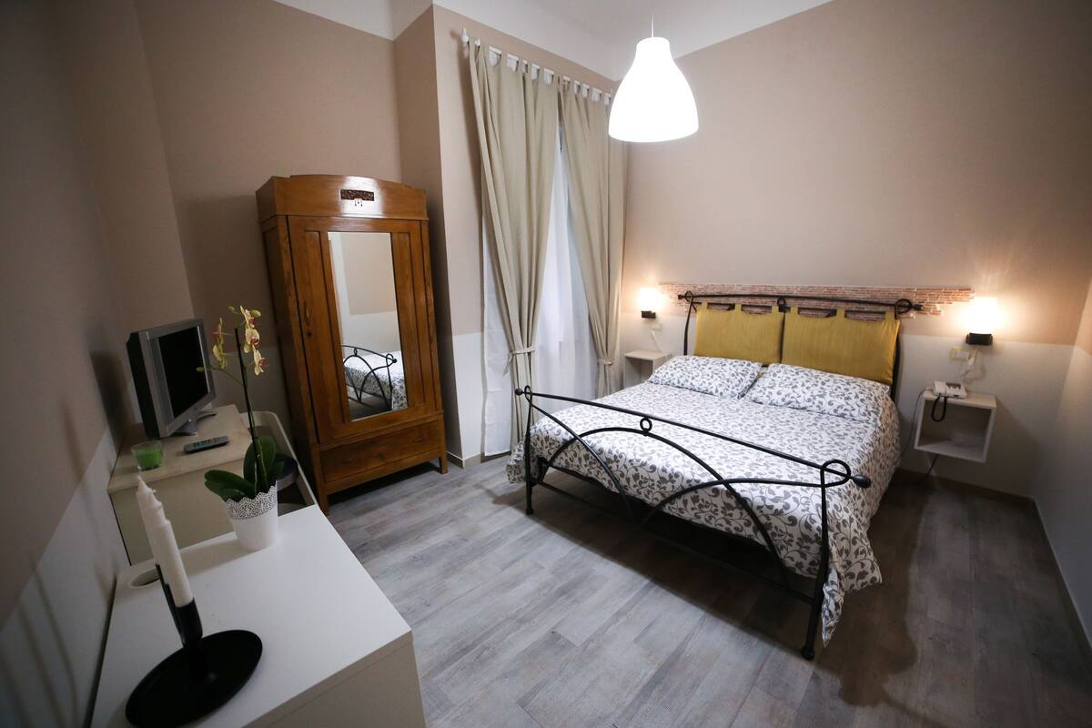 A bedroom with a bed, bedside lamps, a mirror, a dresser, and a desk with a computer and a plant.