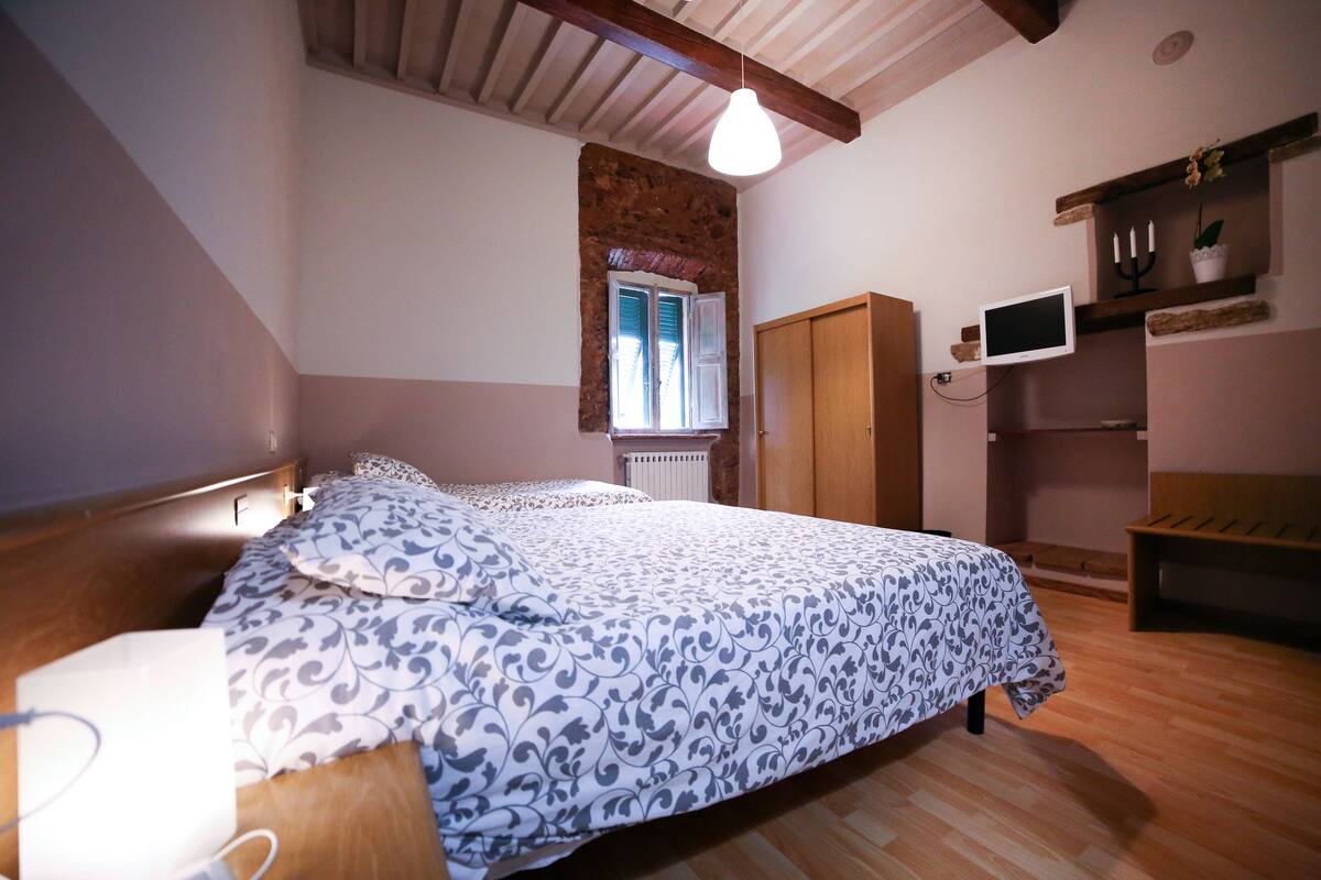 A bedroom with a bed, wooden headboard, a stone wall, a window with shutters, a television on a shelf, and a bench.