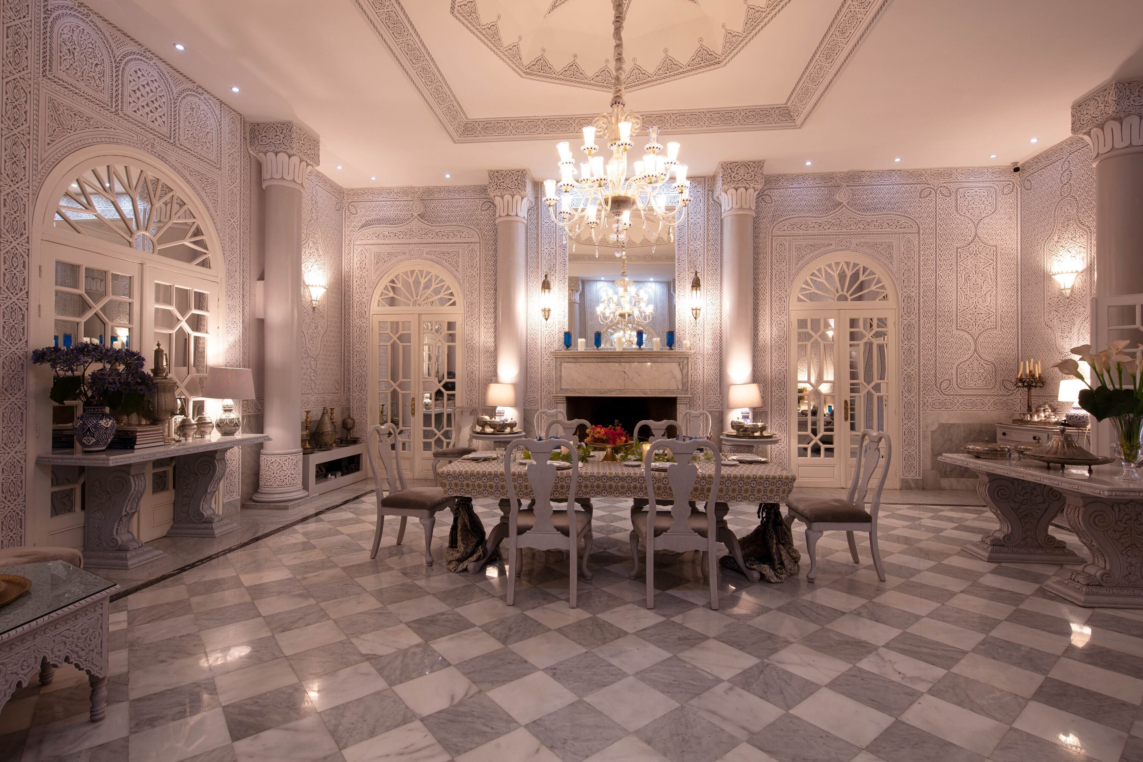 A luxurious dining room with a checkered marble floor, ornate wall designs, a chandelier, and a central fireplace.