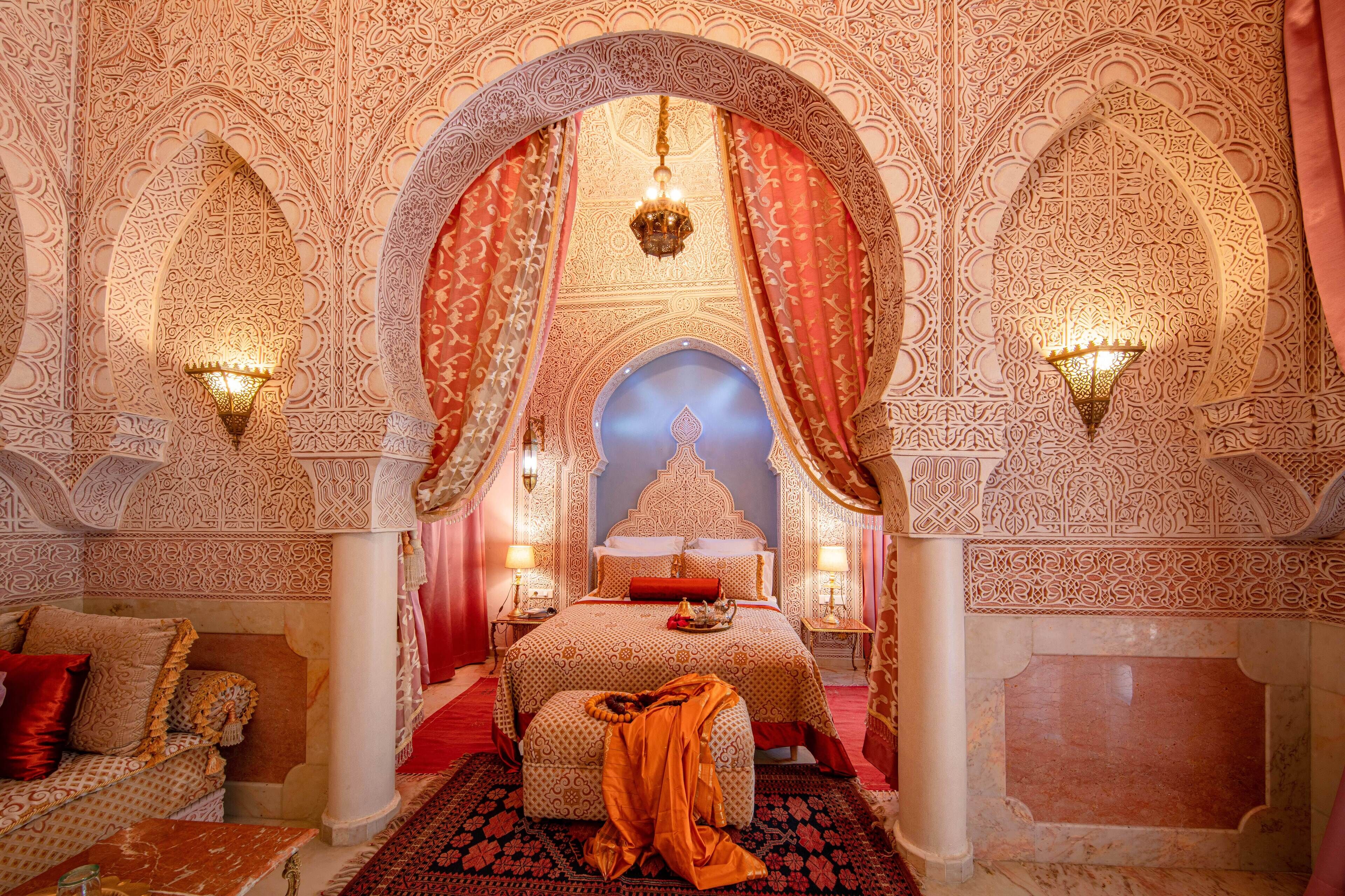 Une pièce richement décorée, ornée de motifs complexes, avec un lit recouvert d’une literie rouge et blanche, et un lustre.