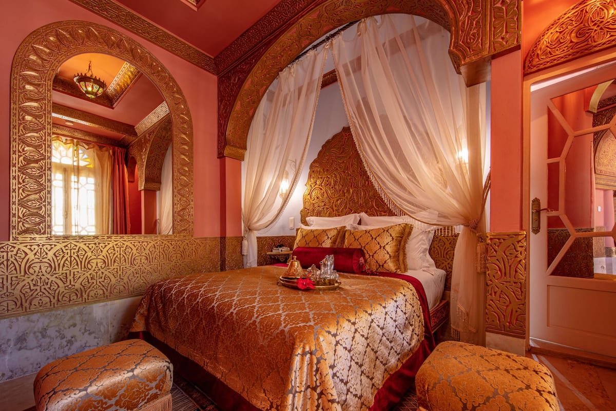 A luxurious bedroom with a four-poster bed, ornate wall designs, and a chandelier.