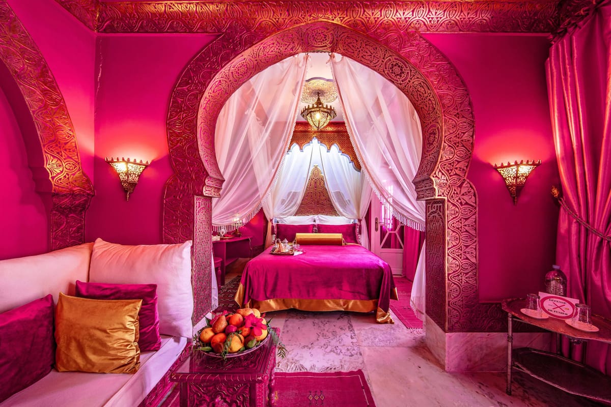 A luxurious room with a large bed, ornate arches, and a canopy bed. There is a sofa with cushions and a table with fruit.