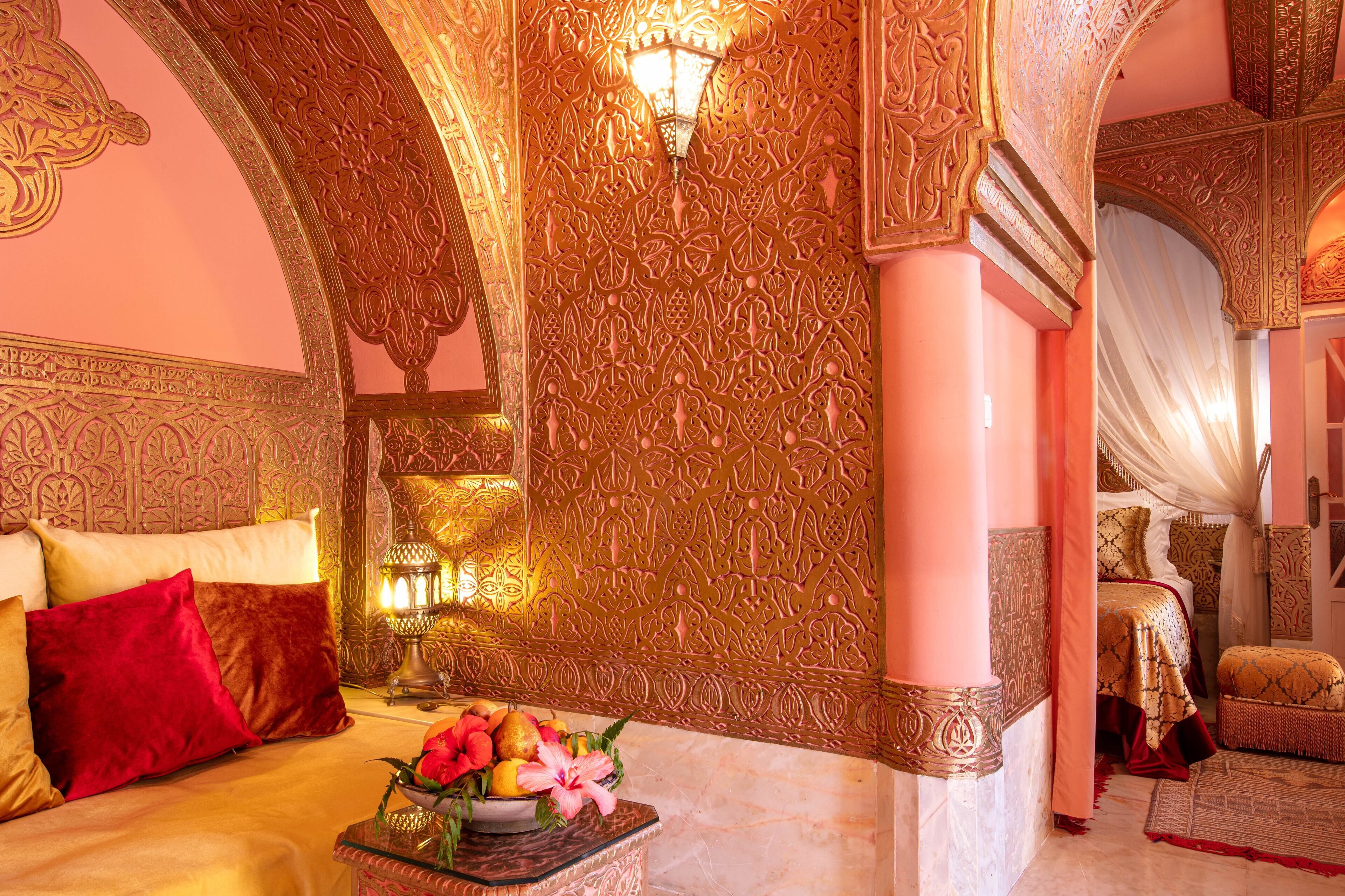 A luxurious room with ornate decor, a bed with red pillows, and a fruit bowl on the nightstand.