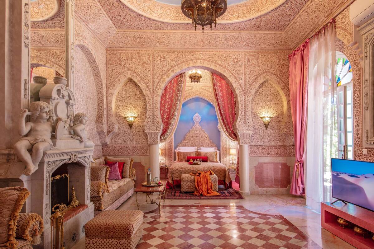A luxurious room with intricate wall designs, a large bed with red and gold accents, a fireplace, and a television.