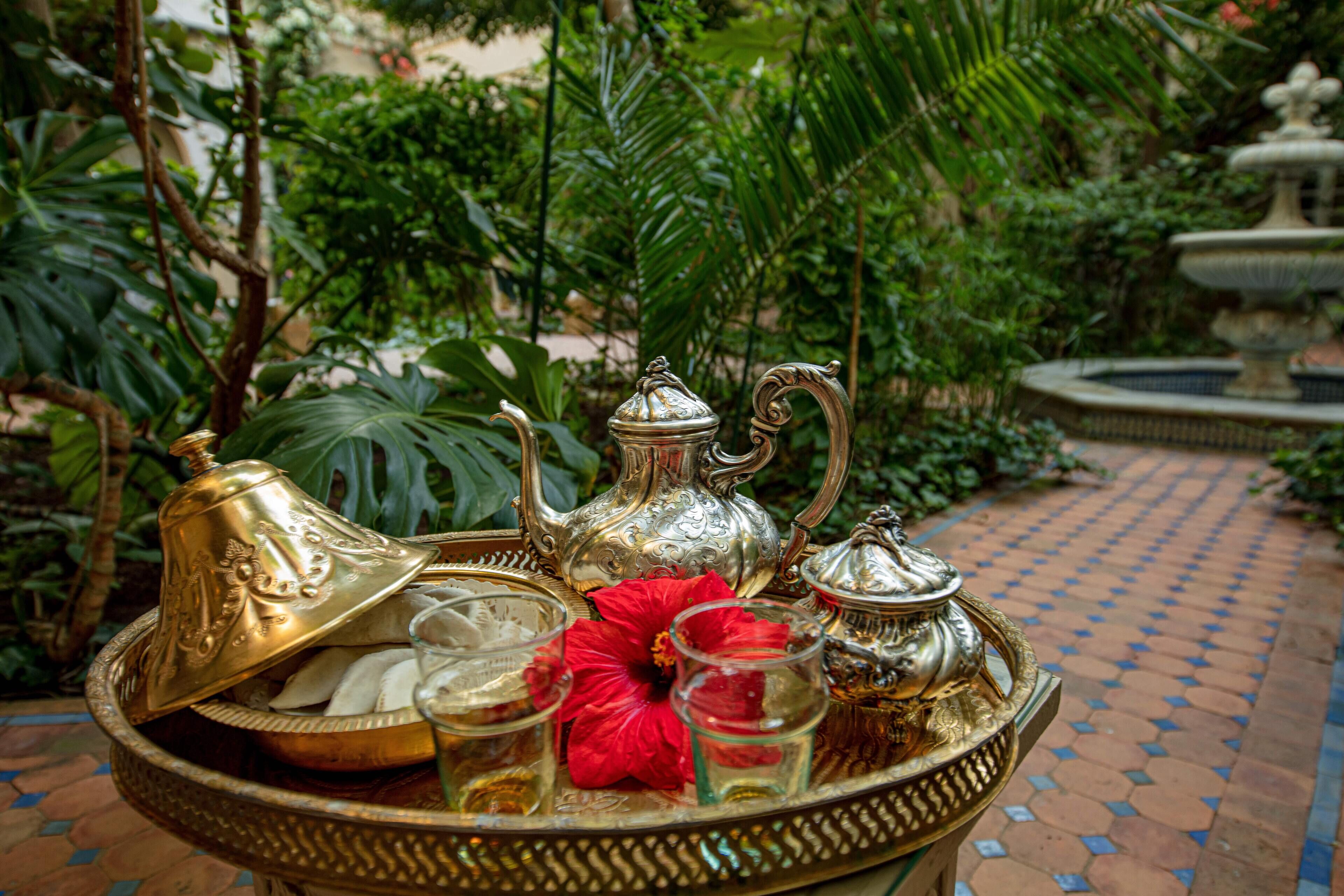 A tray with a brass teapot, sugar bowl, and glasses of tea, set on a tiled floor with a fountain and greenery in the background.