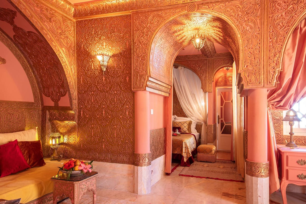 An ornate room with intricate wall carvings, a bed with a red pillow, and a nightstand with a lamp.
