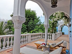 Un área de estar al aire libre con techo, balaustradas blancas, una mesa dispuesta con platos, un farol y vistas a la vegetación y un edificio al fondo.