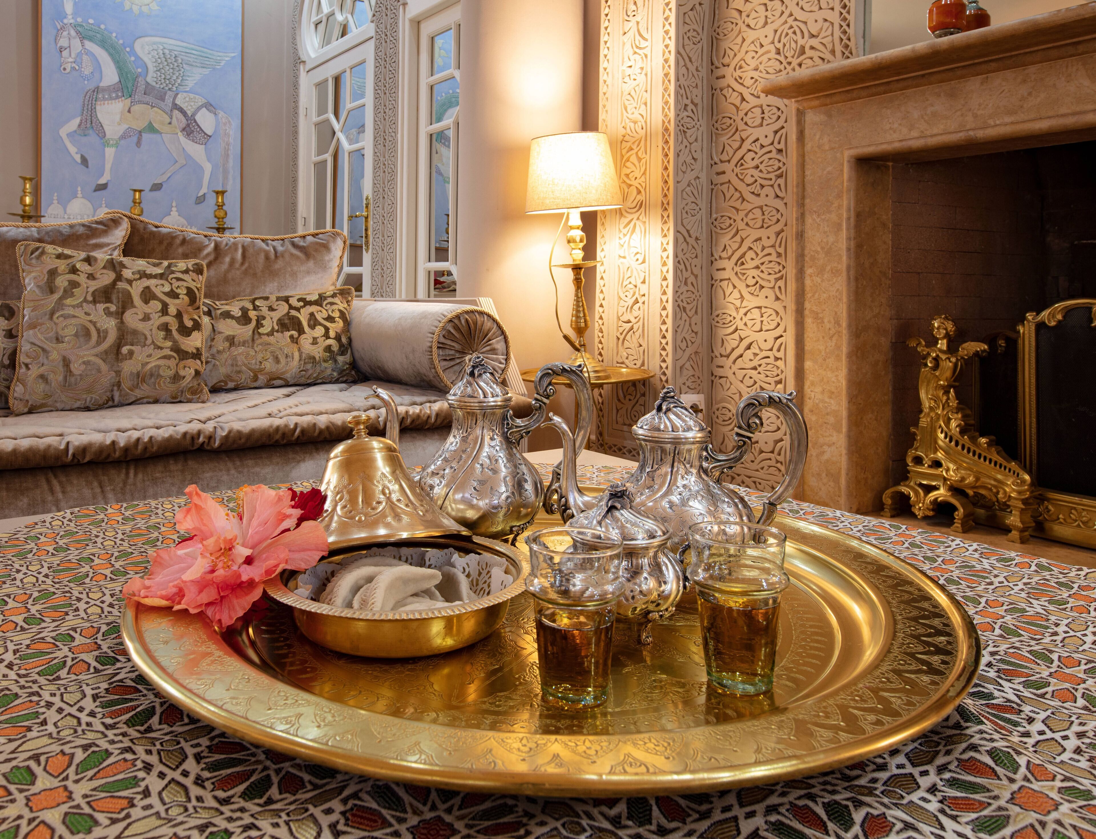 Decorado tradicional marroquí con elegantes juegos de té de plata sobre una bandeja, un sofá con cojines estampados y una pared decorativa con diseños intrincados.