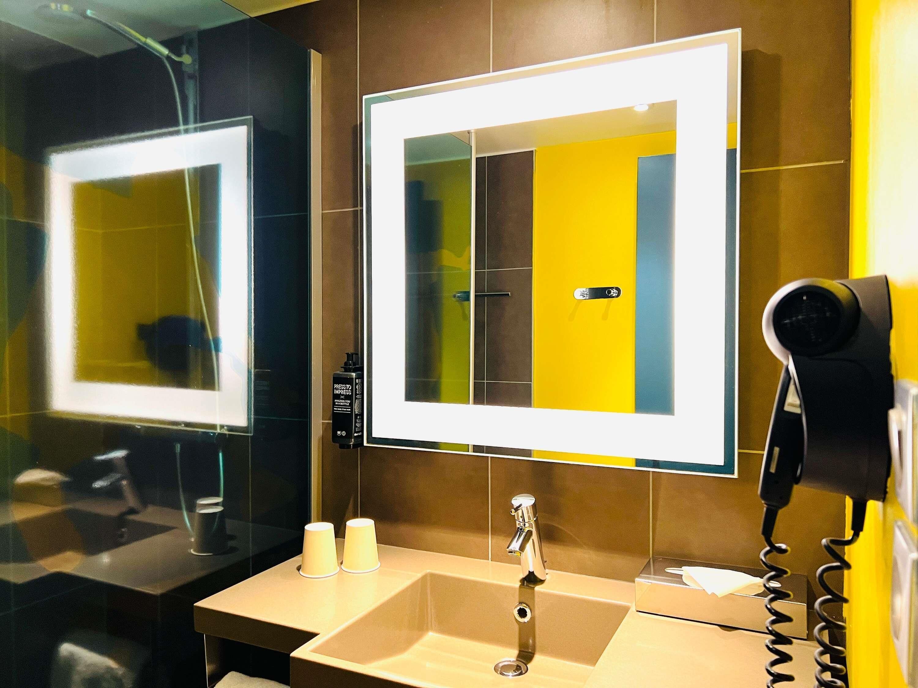 A bathroom with a mirror, a sink, a hairdryer, and a bottle of shampoo.
