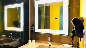 A bathroom with a mirror, a sink, a hairdryer, and a bottle of shampoo.