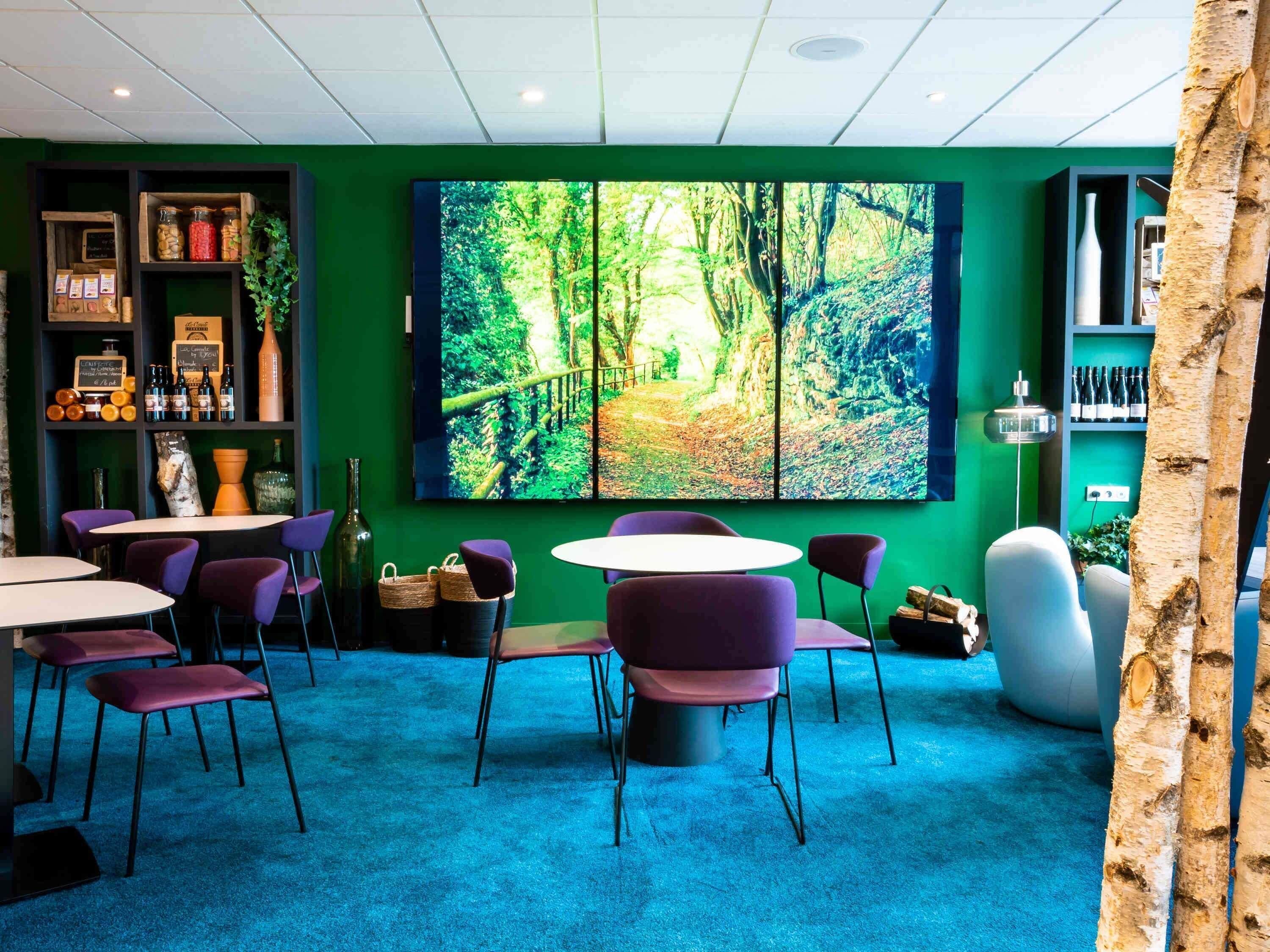 A modern interior with green walls, a large forest-themed painting, purple chairs, and round tables.