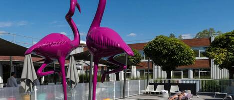 A swimming pool with two pink flamingo sculptures, a covered seating area, and a building with a wooden facade in the background.