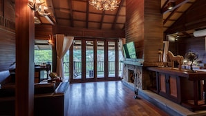 A spacious room with a wooden ceiling, a chandelier, a fireplace, and a large window overlooking greenery.