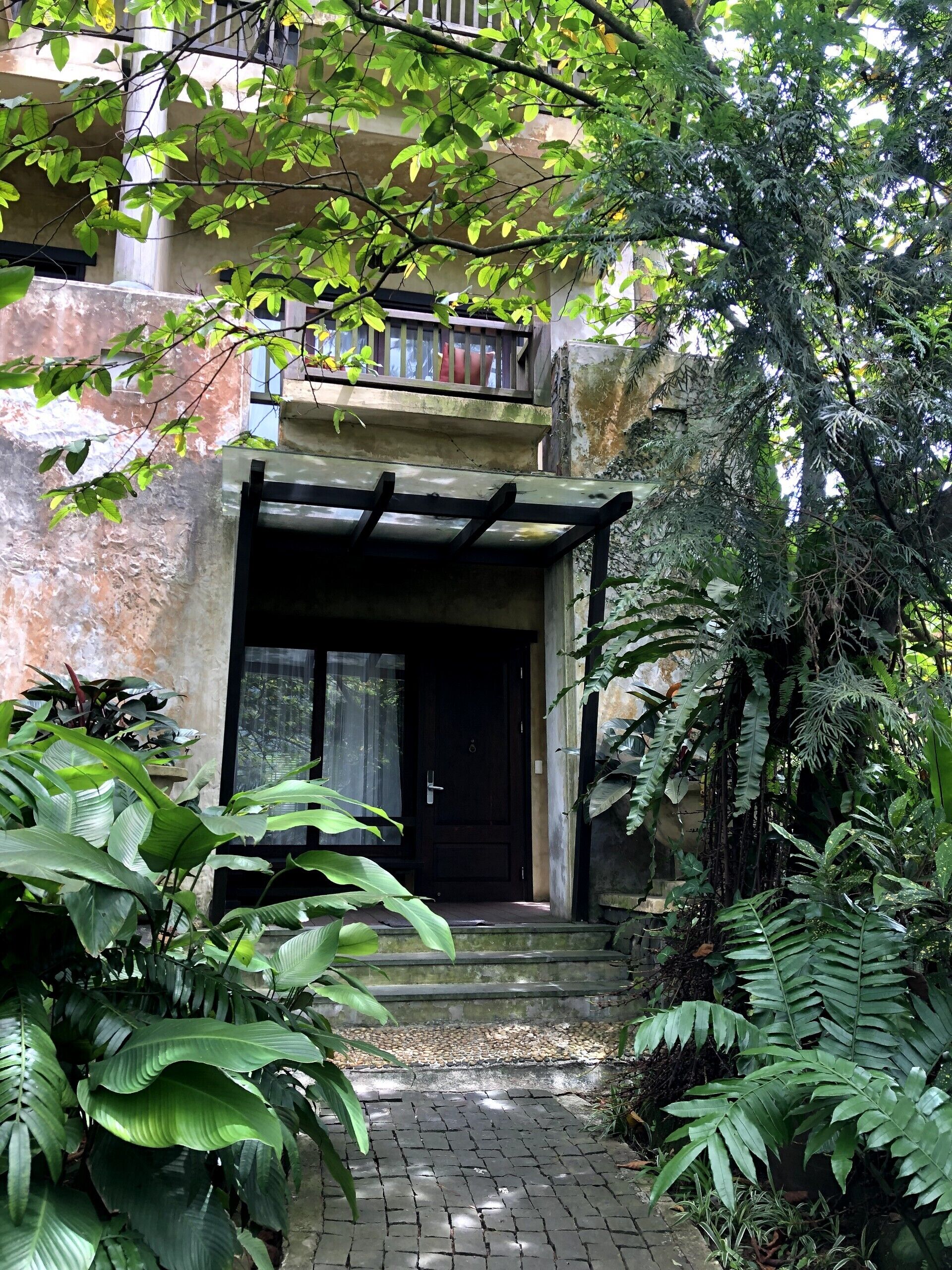 A building with a dark entrance door, a small porch with a wooden railing, and lush greenery surrounding the entrance.