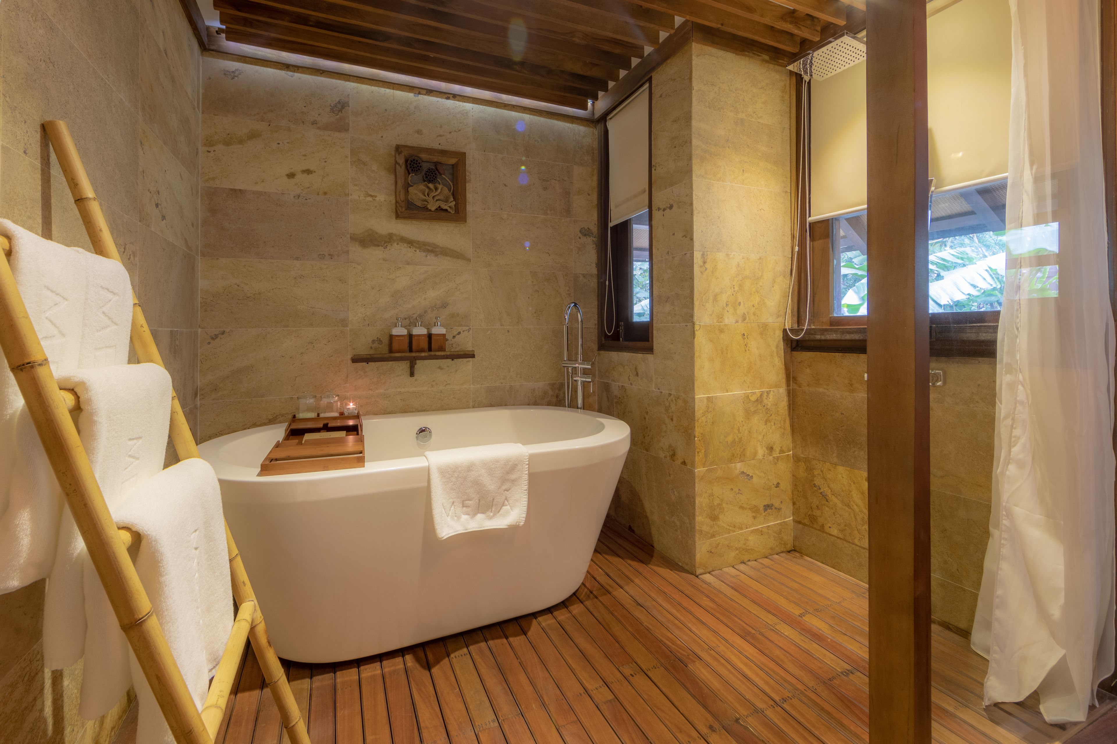 A modern bathroom with a freestanding bathtub, wooden flooring, stone walls, and a window with a view.