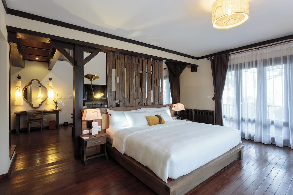 Une chambre spacieuse avec un grand lit, une tête de lit en bois et un plancher en bois. On y trouve deux tables de chevet avec des lampes, un miroir et une fenêtre ornée de rideaux légers.