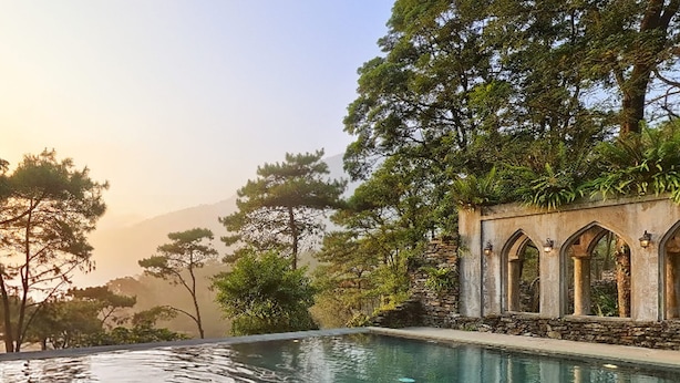 A pool with a stone wall and arched windows, surrounded by trees and a mountainous backdrop.