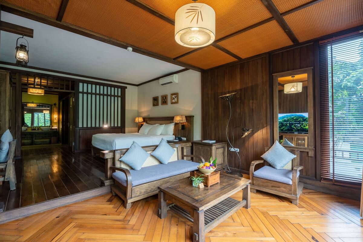 Une chambre au décor traditionnel, comprenant un lit, deux canapés, une table basse en bois et une grande fenêtre avec des stores.