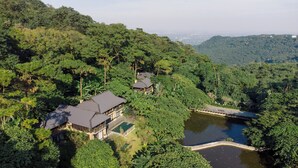 A secluded resort with multiple buildings surrounded by dense forest, a small lake, and a bridge connecting the structures.
