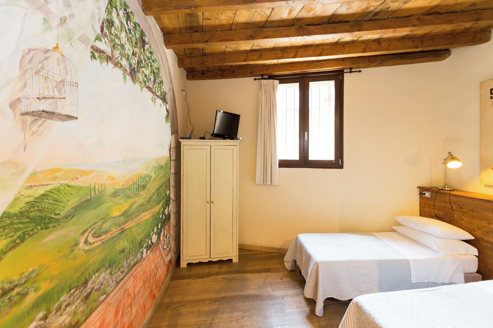 A room with two beds, a wooden ceiling, a mural on the wall, a cabinet, and a window with curtains.