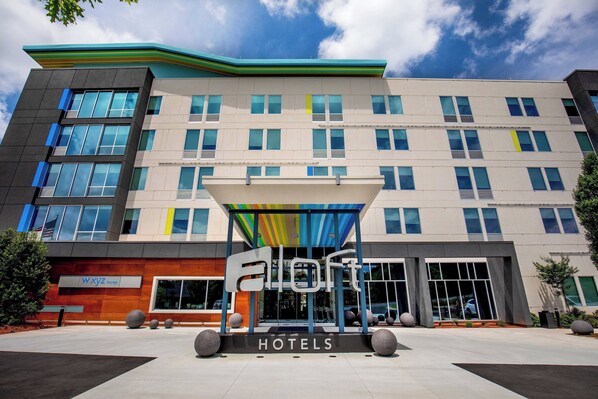 Un edificio moderno de hotel con un llamativo letrero de 'ELoft Hotels', grandes ventanales y un área de entrada cubierta.