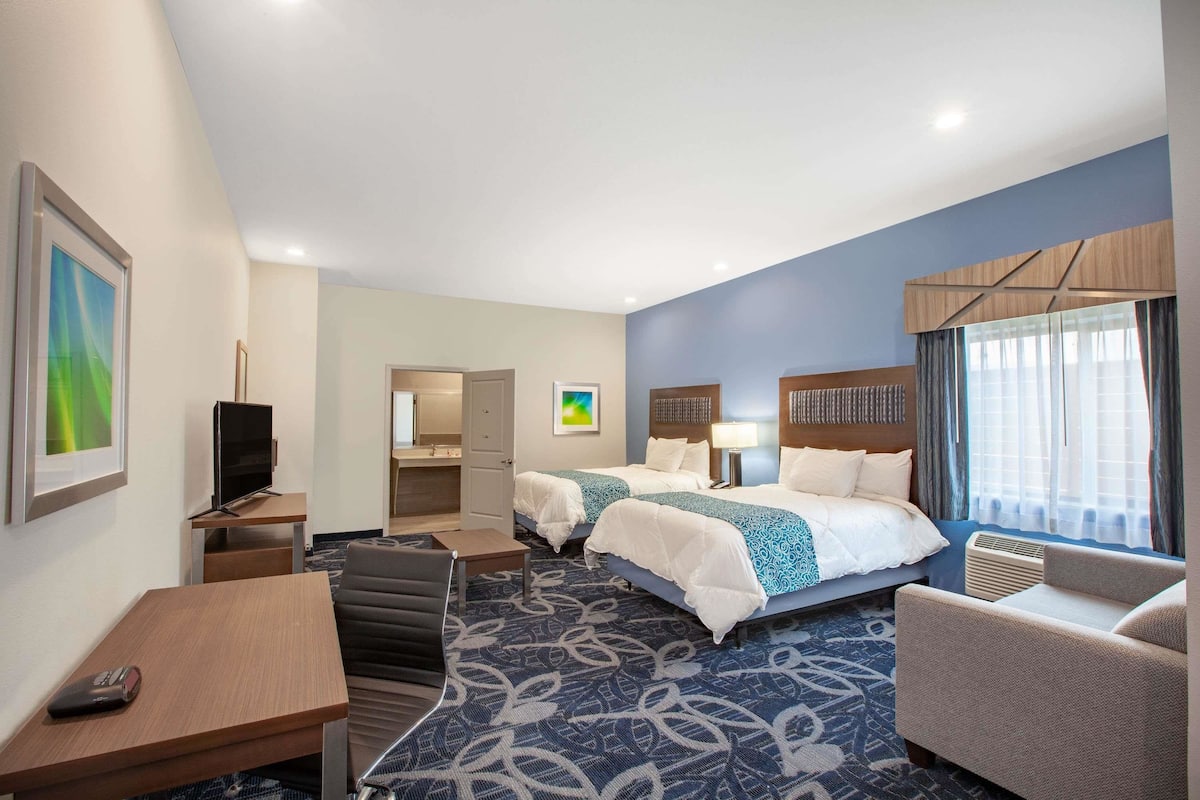 A hotel room with two beds, a desk, a TV, a chair, and a large window with curtains.