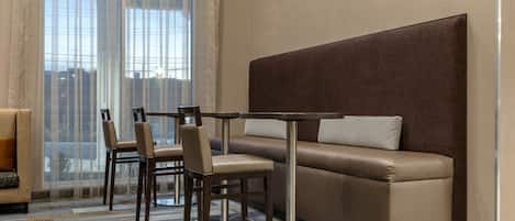 A modern hotel room with a sofa, two chairs, a small table, and a large window with sheer curtains.