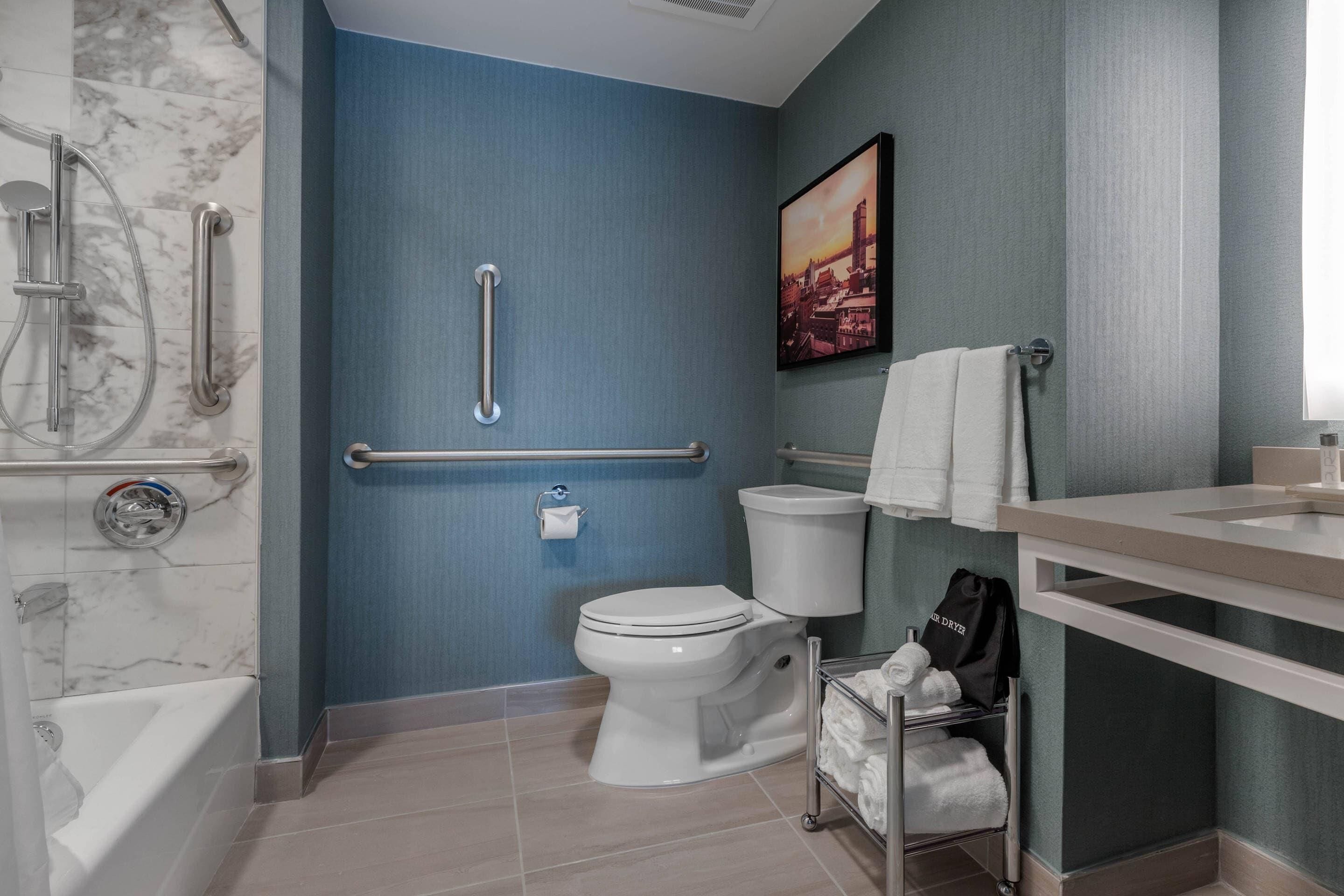 Une salle de bain avec des toilettes blanches, une barre d’appui, un porte-serviettes et un espace douche.