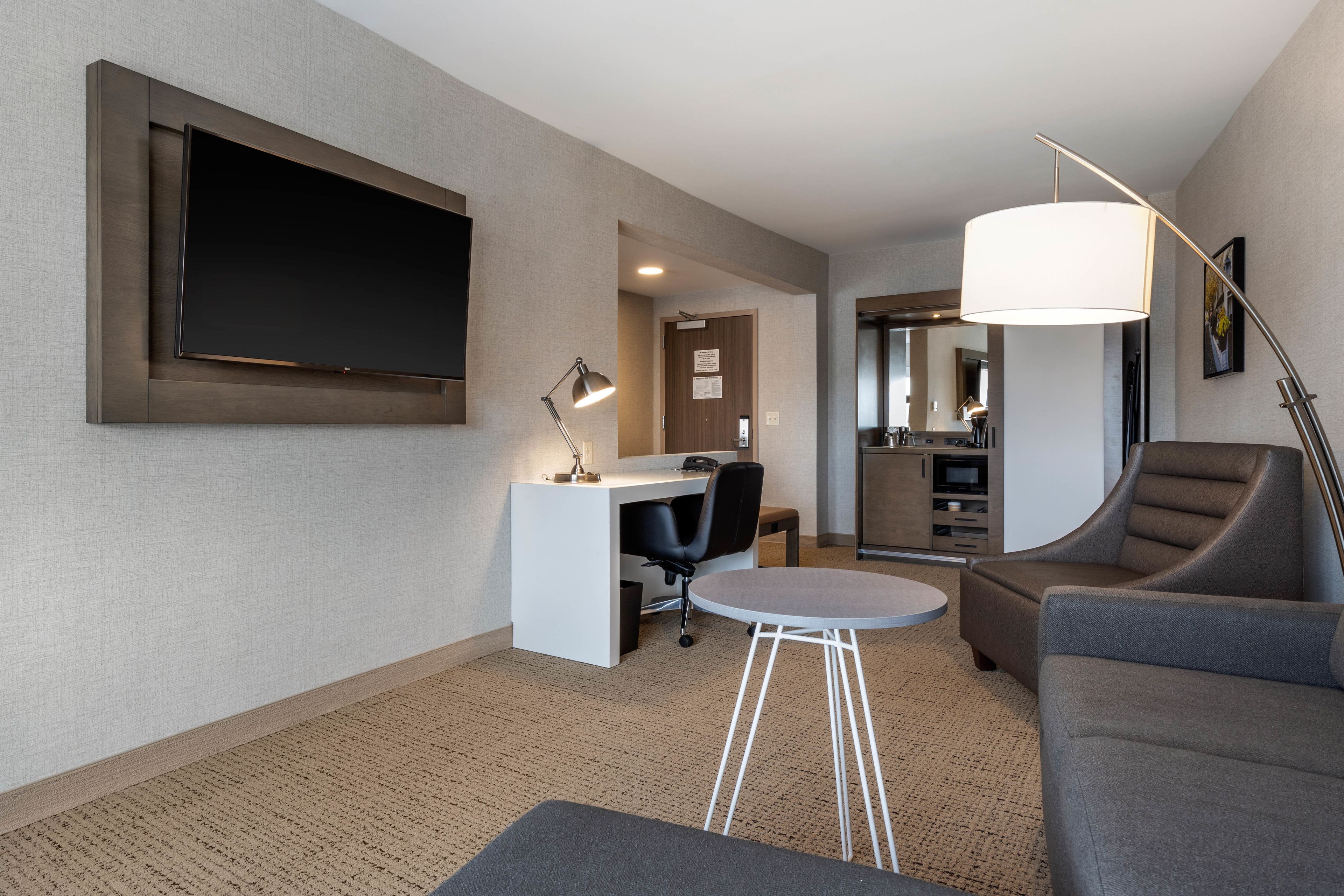 A modern hotel room with a flat-screen TV, a desk with a chair, a round coffee table, a sofa, and a large floor lamp.