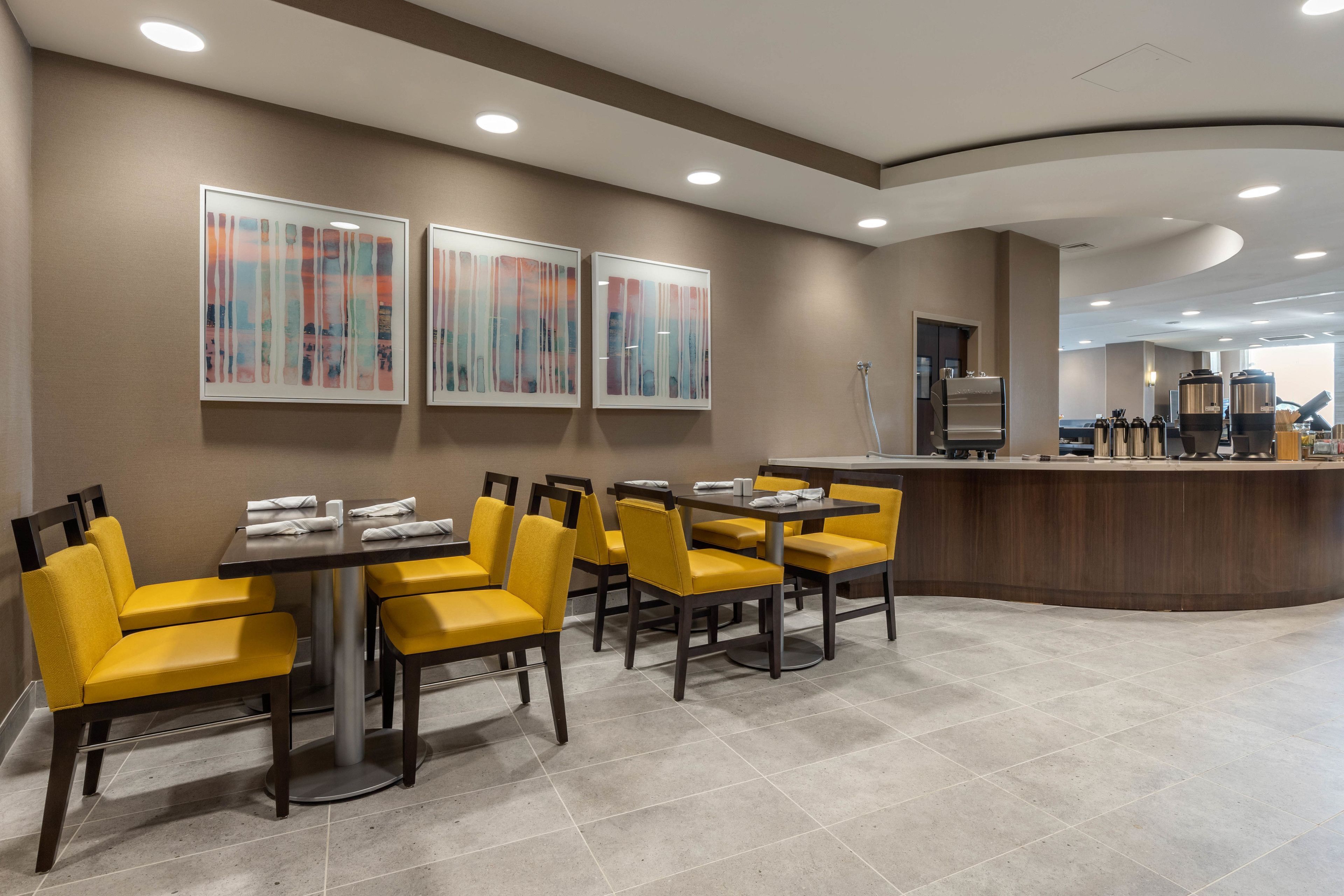 A modern dining area with yellow chairs and tables, a counter with coffee machines, and abstract art on the walls.