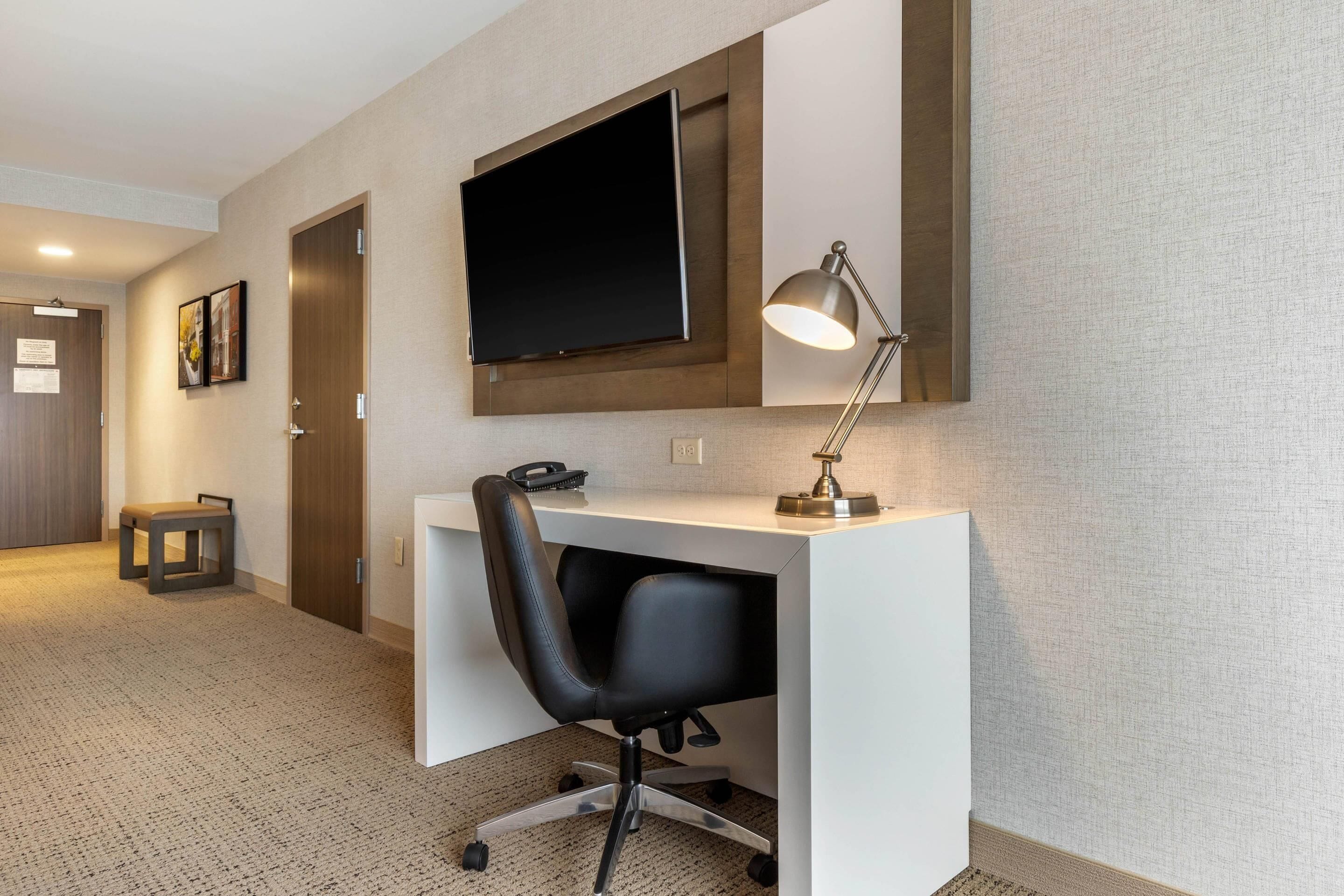 A hotel room with a desk, chair, TV, and lamp.