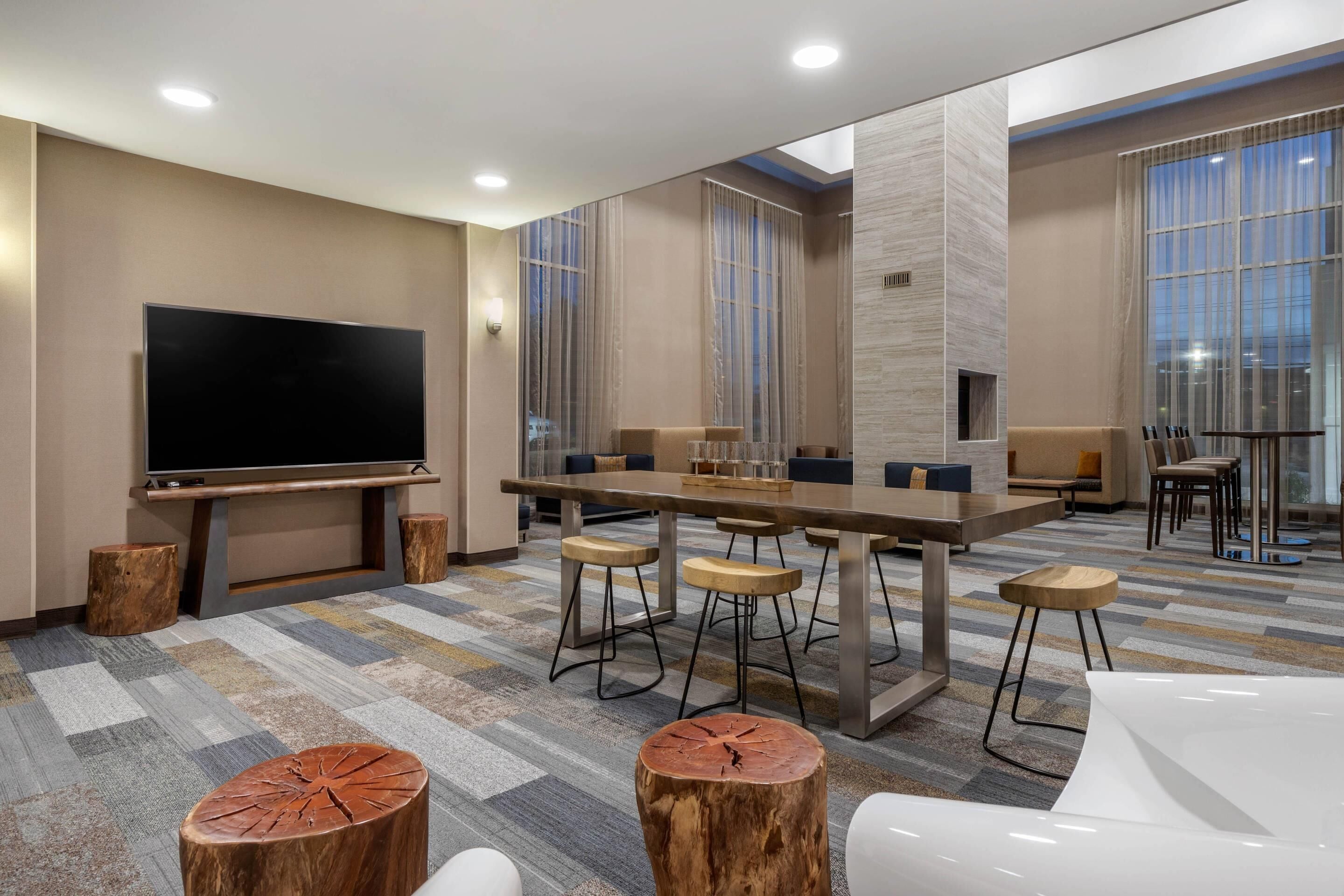 Ein modernes Hotelzimmer mit einem großen Flachbildfernseher, einem langen Esstisch mit Hockern und einer Sitzecke mit Sofas und Sesseln.