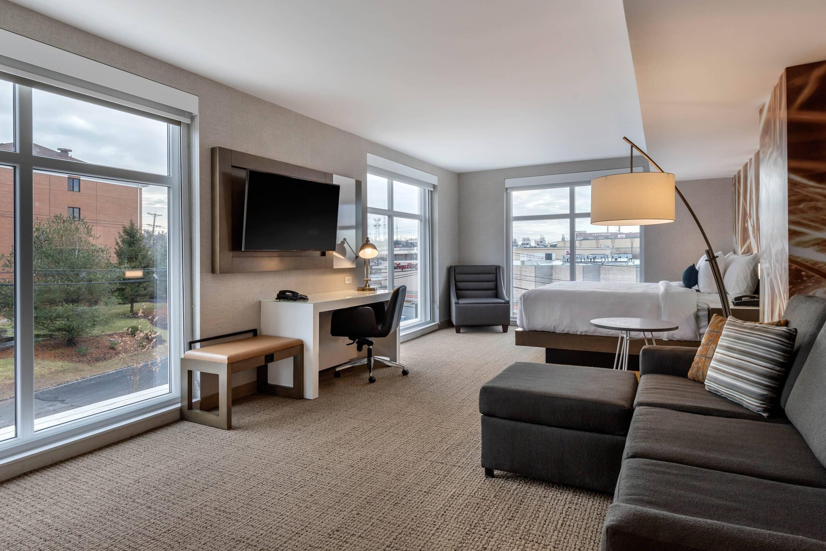 A hotel room with a bed, a desk with a chair, a sofa, a TV mounted on the wall, and large windows offering a view of the city.