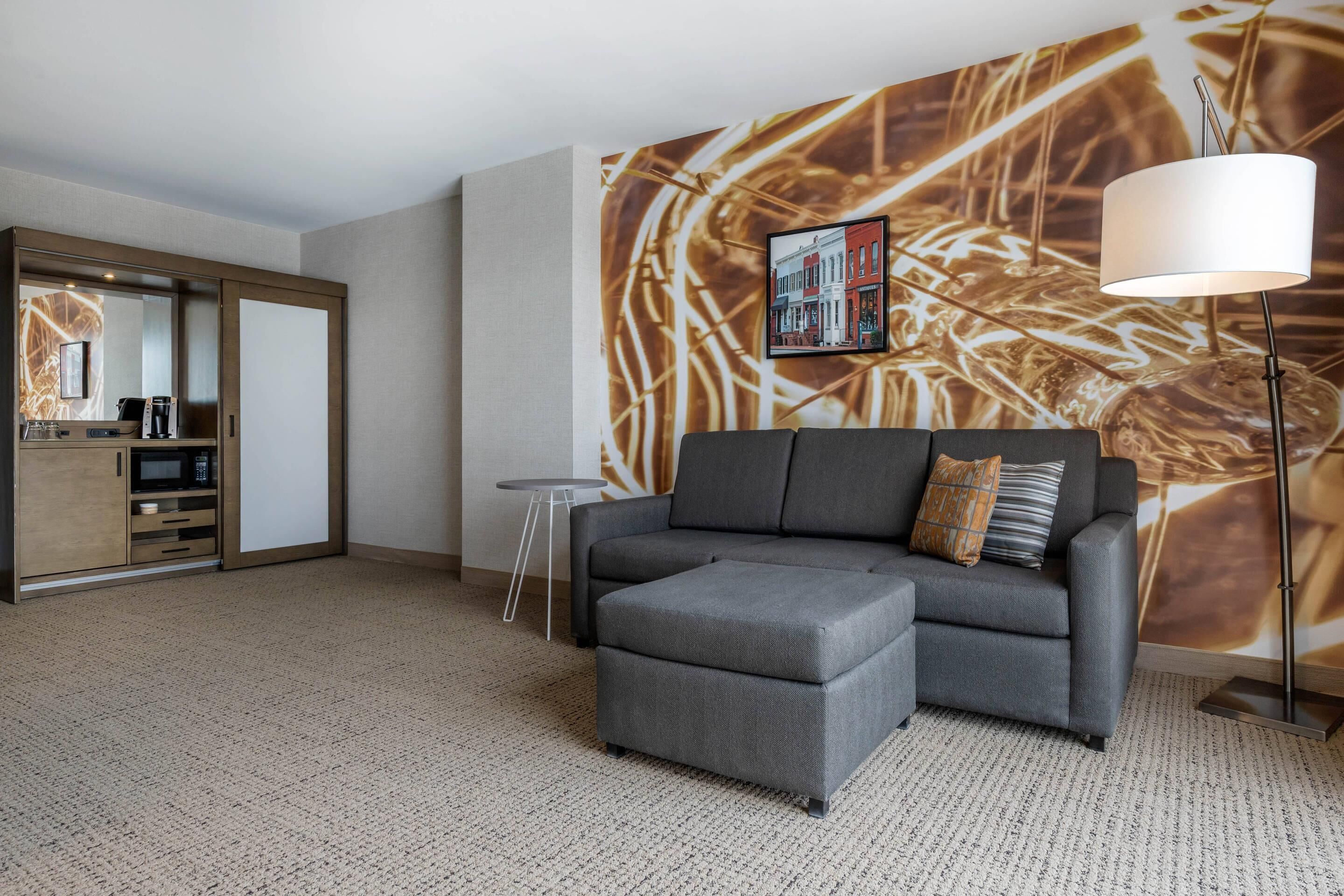 A modern hotel room with a grey sofa, a small table, a floor lamp, and a large abstract wall mural.