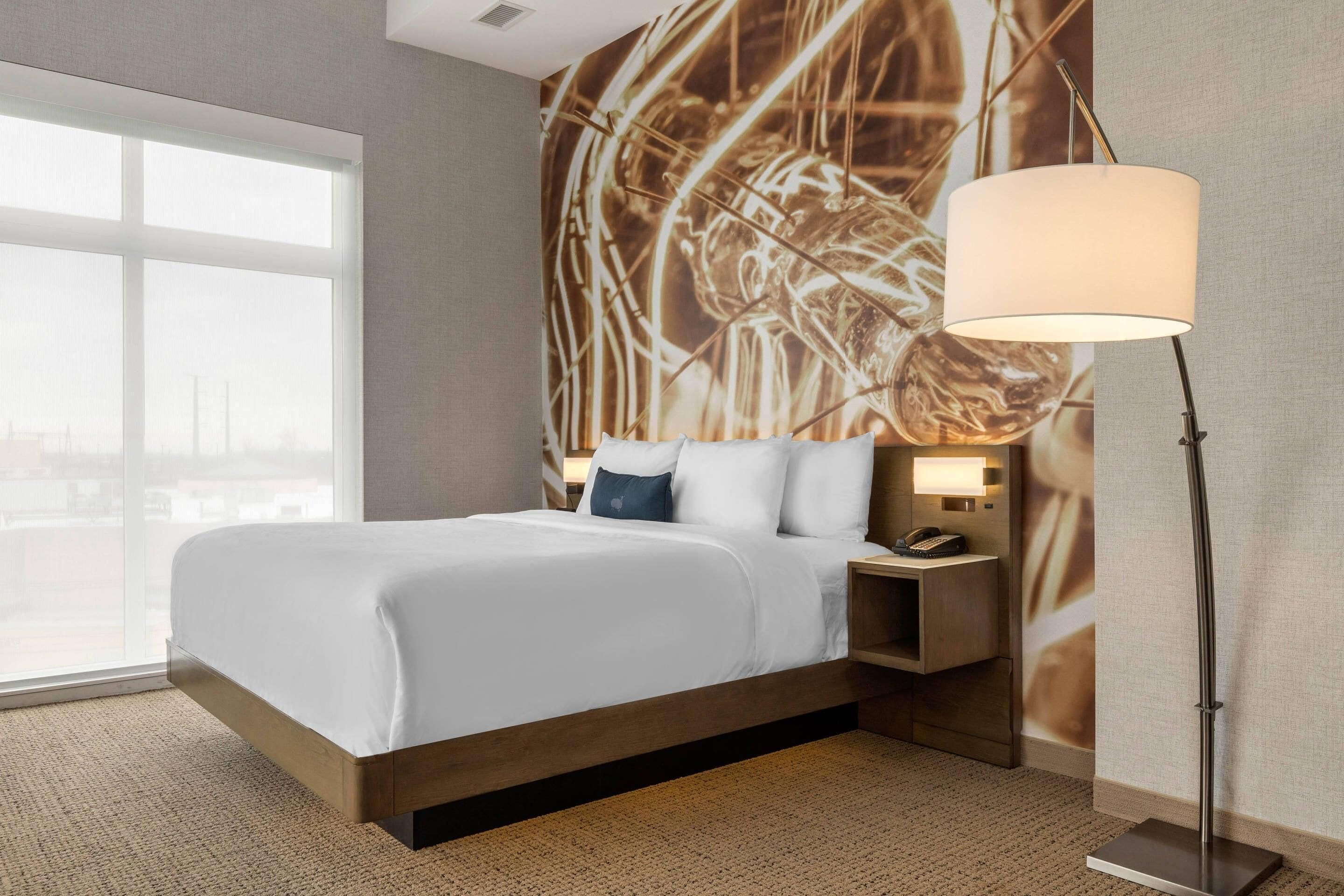 A hotel room with a large bed, a nightstand with a lamp, and a wall with a large abstract painting.
