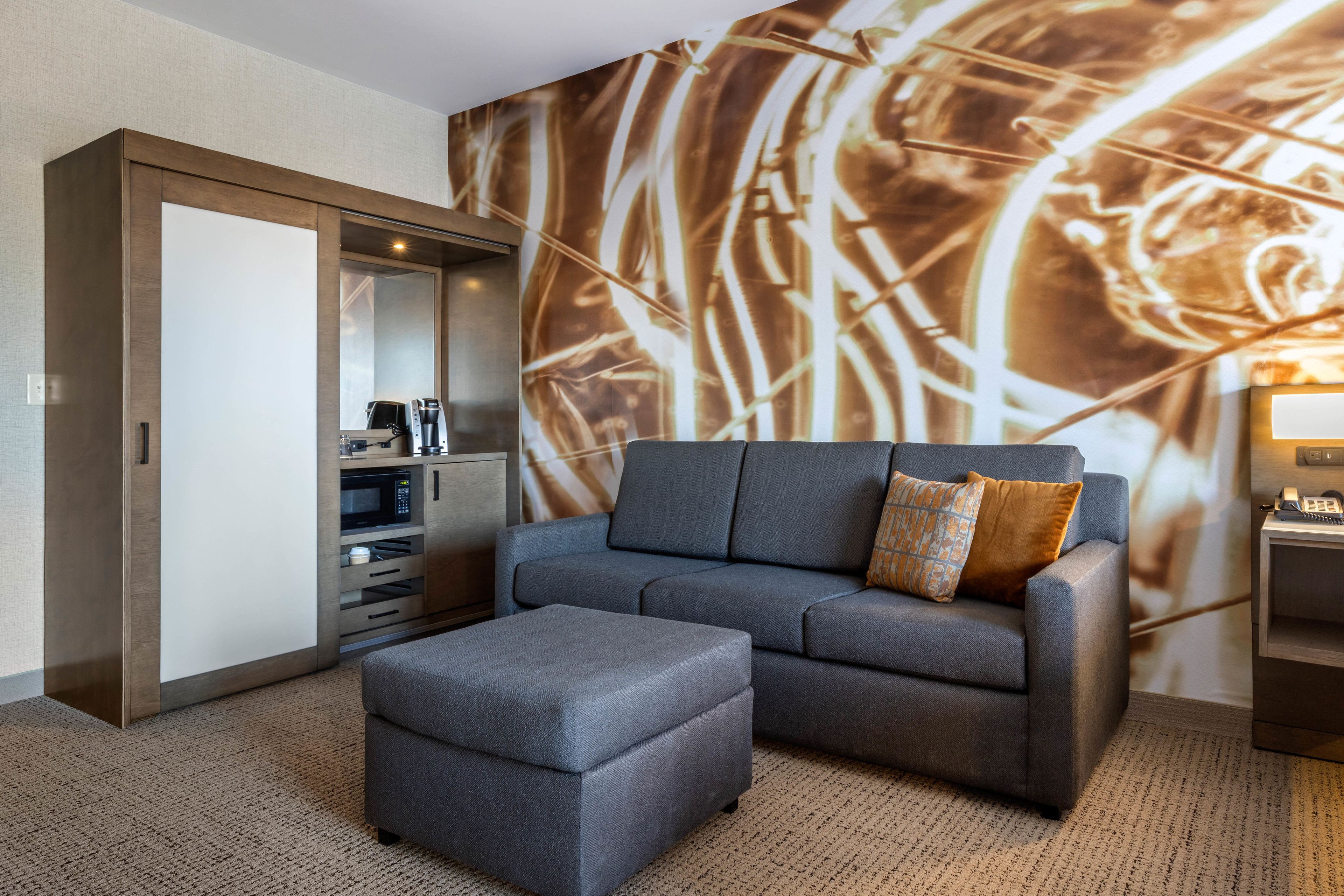 A modern hotel room with a grey sofa, a matching ottoman, a microwave, and a coffee machine.