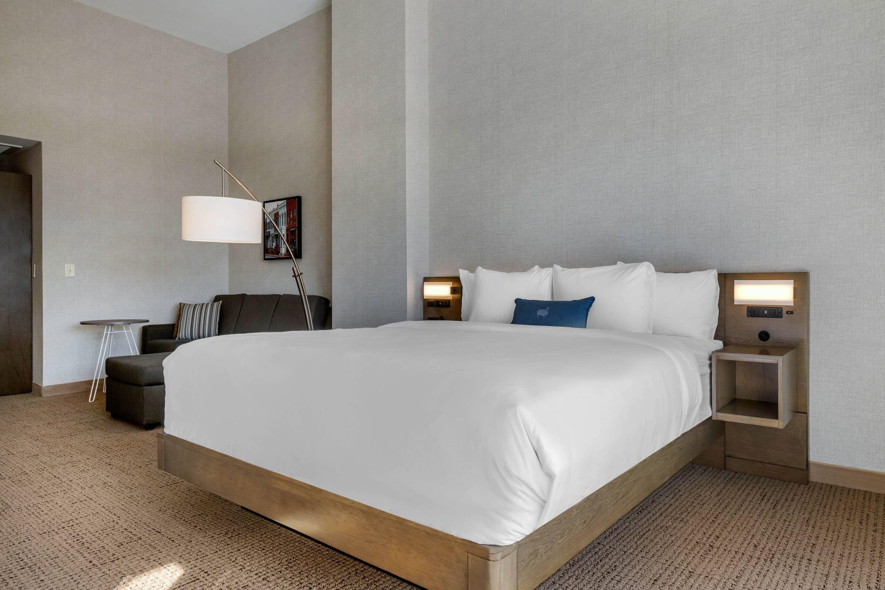 Habitación de hotel con una cama grande, un escritorio, una silla y una mesita de noche con lámpara.