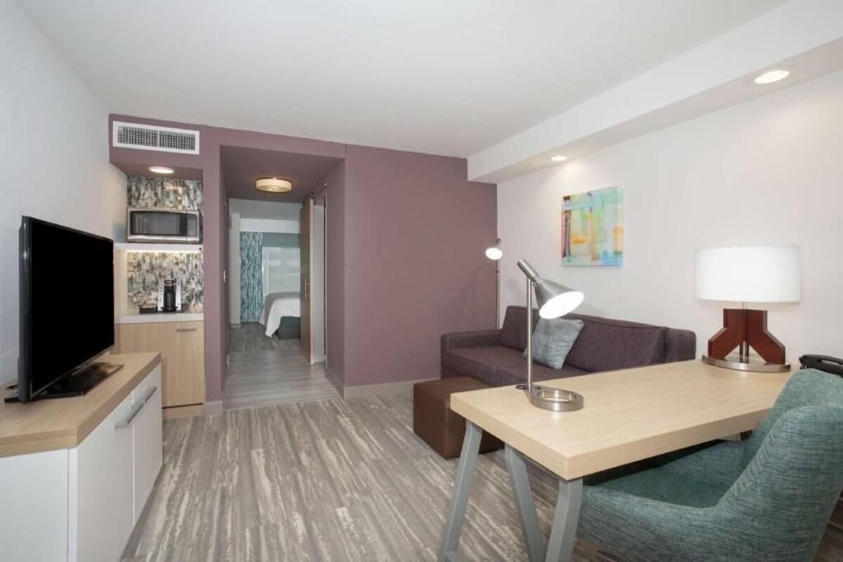 Habitación de hotel moderna con televisión, kitchenette, comedor y vistas al baño.