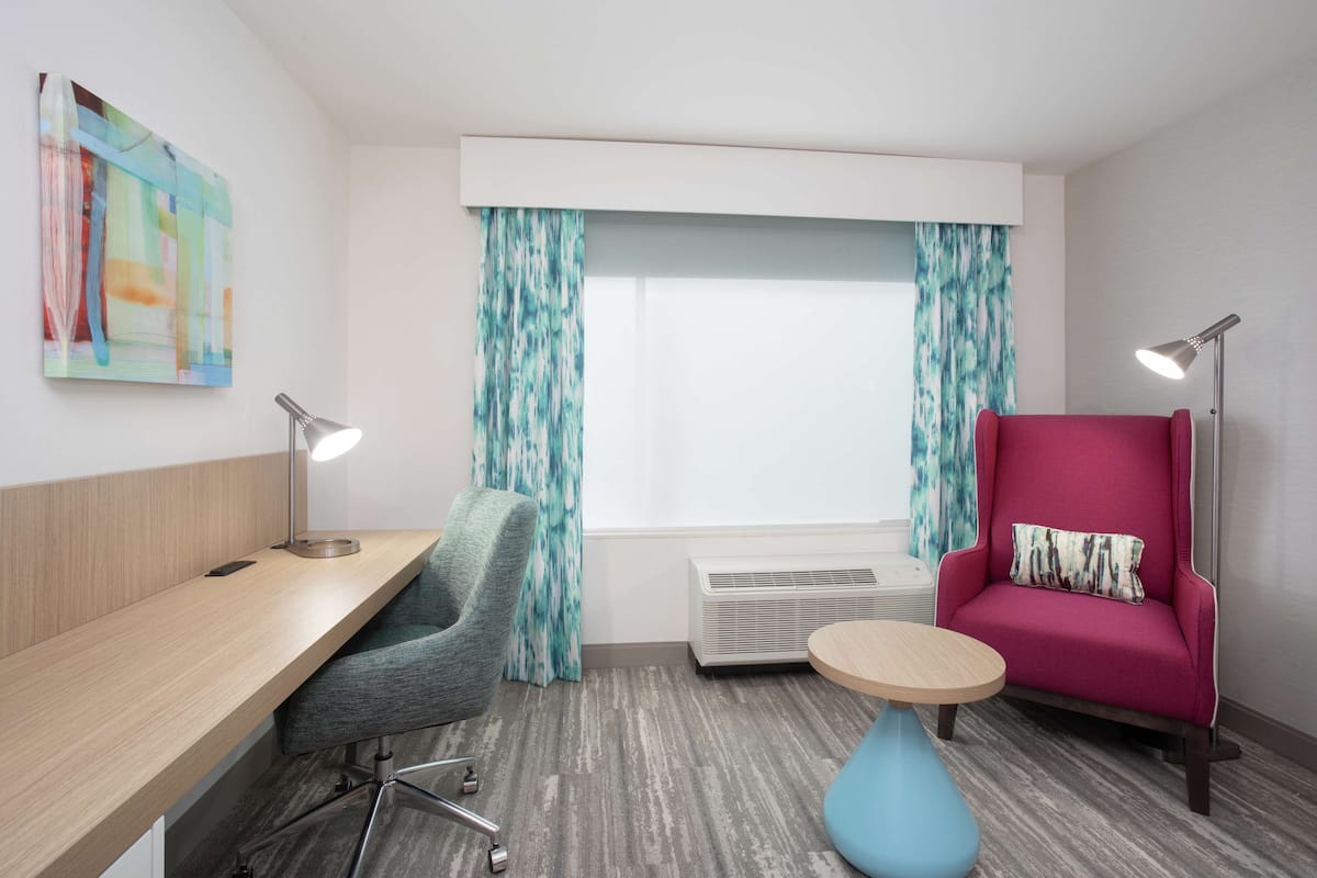 Una habitación de hotel moderna con un escritorio de madera, una silla de oficina gris, un sofá rosa, una mesa redonda azul y un ventanal con cortinas.