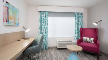 A modern hotel room with a wooden desk, a grey office chair, a pink sofa, a round blue table, and a large window with curtains.