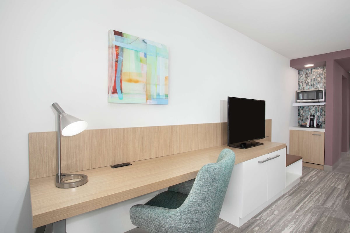 Una habitación moderna con un escritorio de madera, una silla verde, un televisor de pantalla plana, una lámpara de escritorio y un cuadro abstracto de colores en la pared.