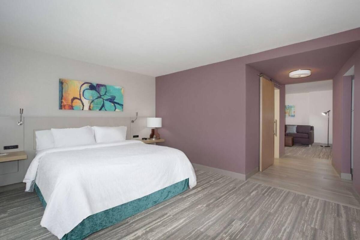 Habitación de hotel con cama, mesita de noche, lámpara, un cuadro en la pared y un pasillo que conduce a otra habitación.