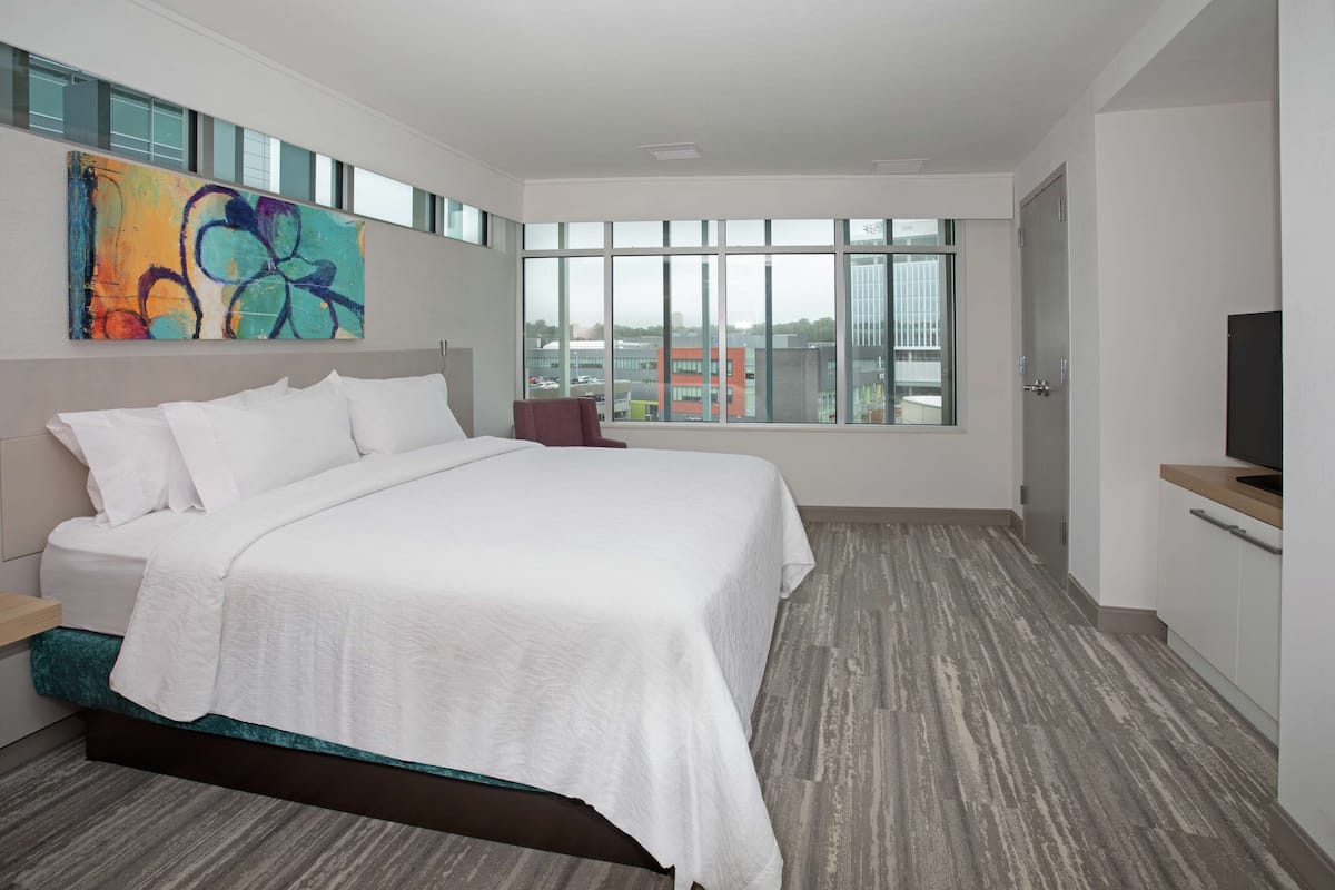 Una habitación de hotel moderna con una cama grande, televisión y vistas a la ciudad a través de amplias ventanas.