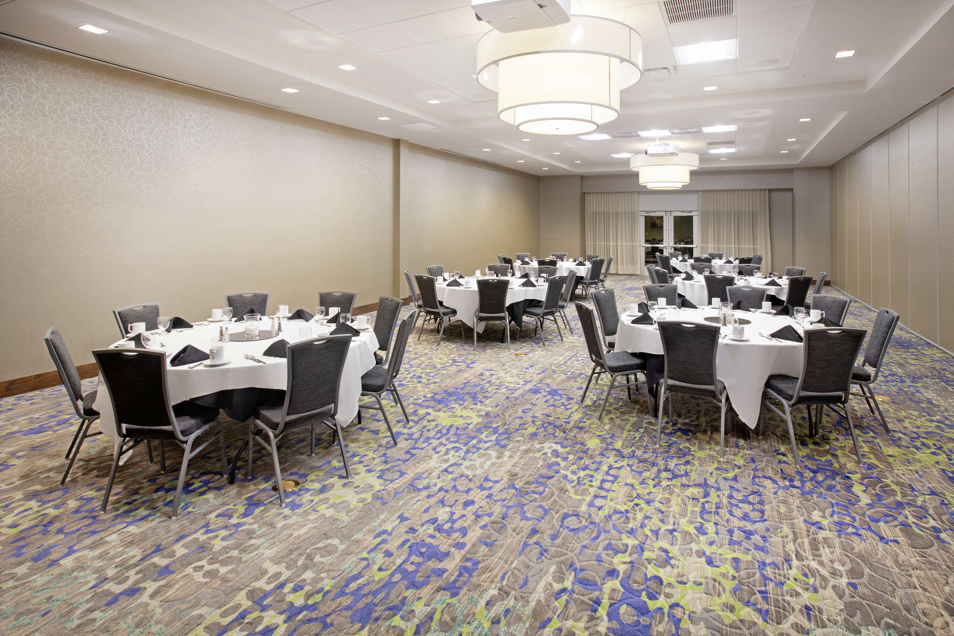 A conference room with multiple tables and chairs, arranged in rows, with a patterned carpet and a plain wall.