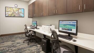 A conference room with two desks, each with a computer, keyboard, and mouse. There are two office chairs, a clock on the wall, and a carpeted floor.