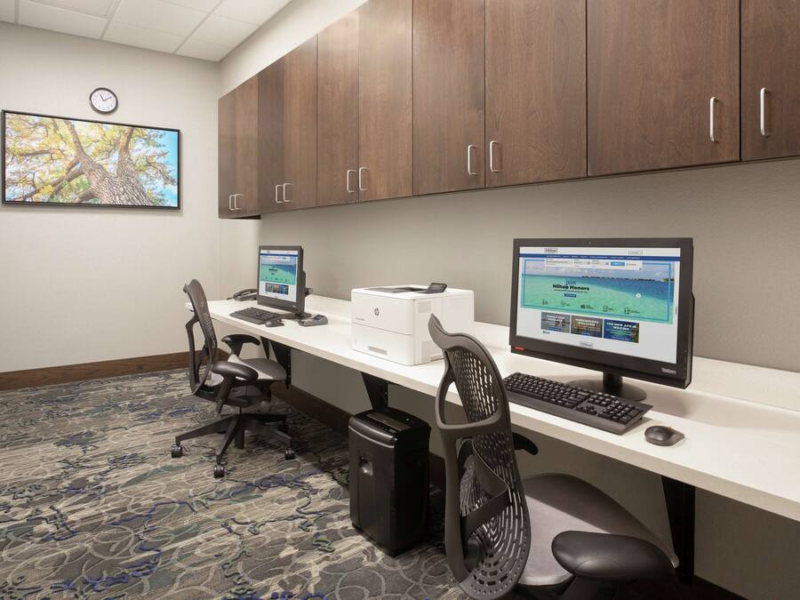 A conference room with two desks, each with a computer, keyboard, and mouse. There are two office chairs, a clock on the wall, and a carpeted floor.