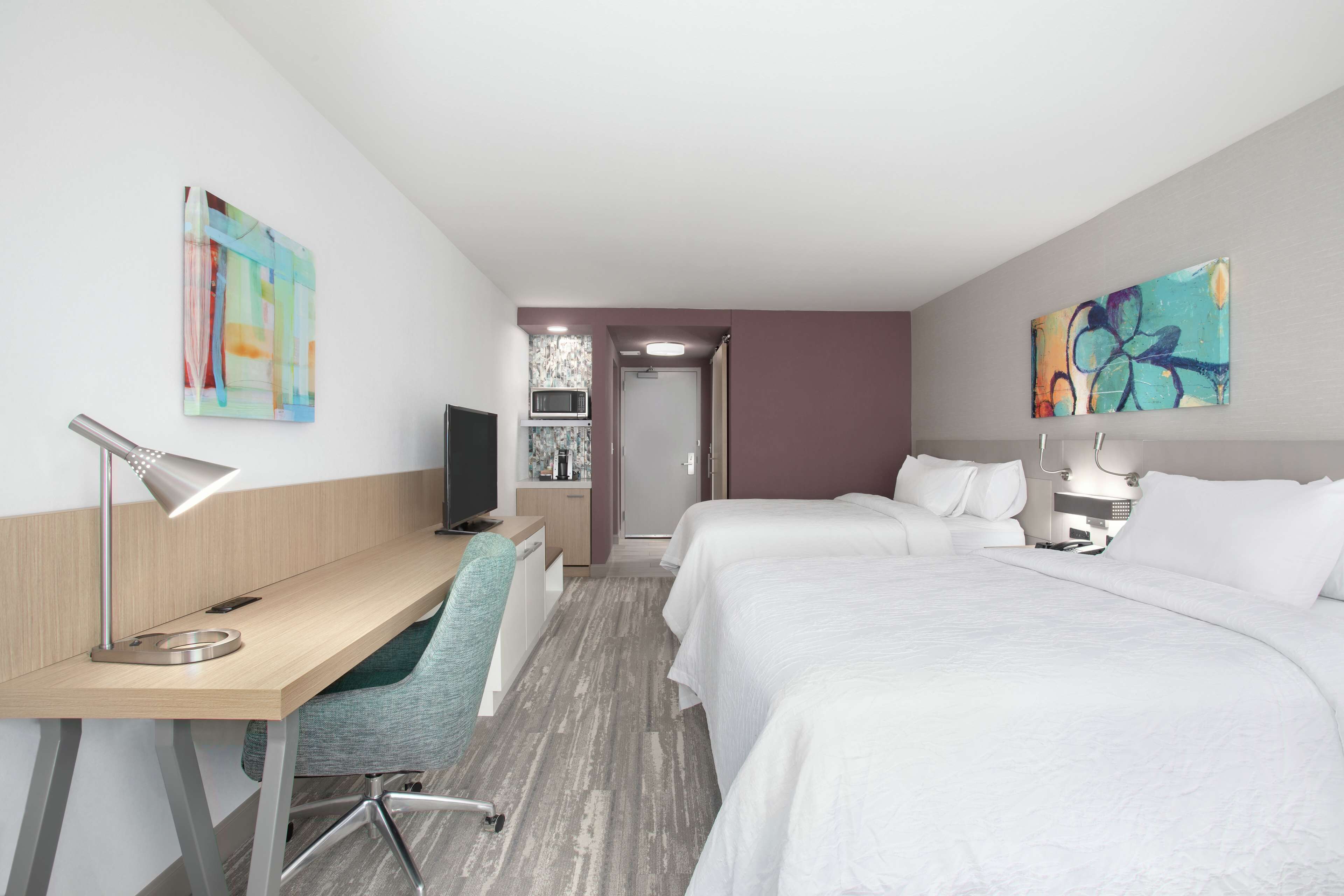 Habitación de hotel con escritorio, dos camas, una silla, televisor y obras de arte en las paredes.