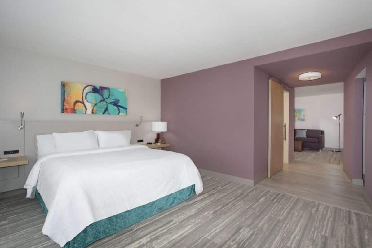 Habitación de hotel con cama, mesita de noche, lámpara, un cuadro en la pared y un pasillo que conduce a otra habitación.