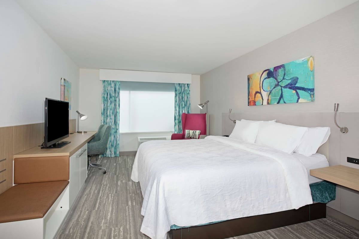 Habitación de hotel con cama, escritorio con televisor, silla, ventana con cortinas y un cuadro en la pared.