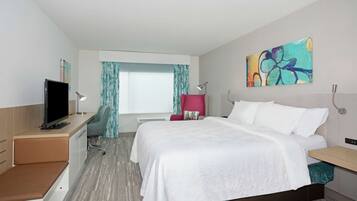 A hotel room with a bed, a desk with a TV, a chair, a window with curtains, and a painting on the wall.